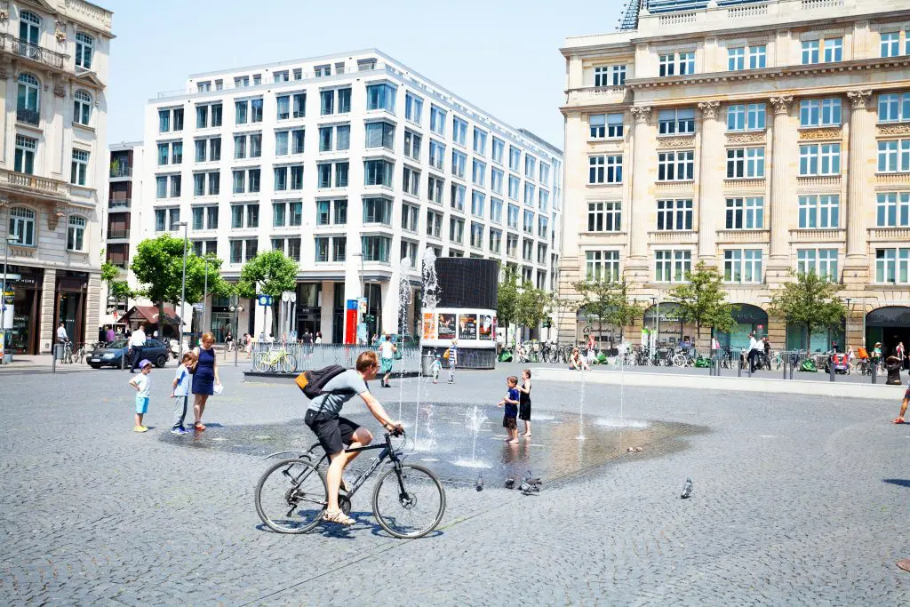 Frankfurt Main, Germany - Capture of people on square Goetheplatz and surrounding office buildings in summer. Some children are playing at fountain. A man is cycling in scene.