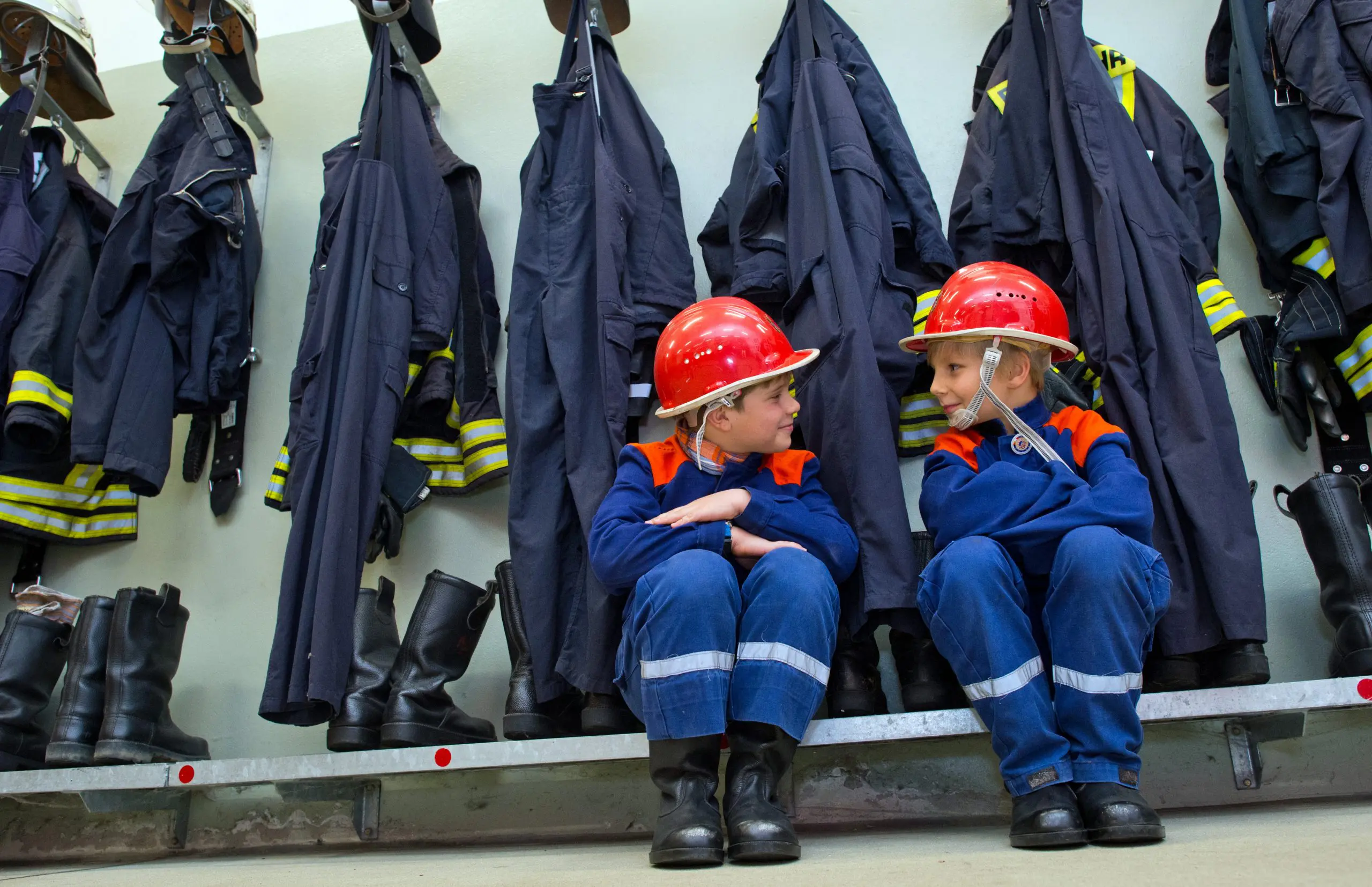 Nachwuchs für die Feuerwehr - zwei Kinder in Feuerwehr-Anzügen sitzen in der Garderobe vor Feuerwehrjacken
