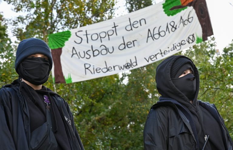 Zwei Aktivistinnen, hinter denen ein Plakat mit der Aufschrift "Stoppt den Ausbau der A661 & A66. Riederwald verteidigen"