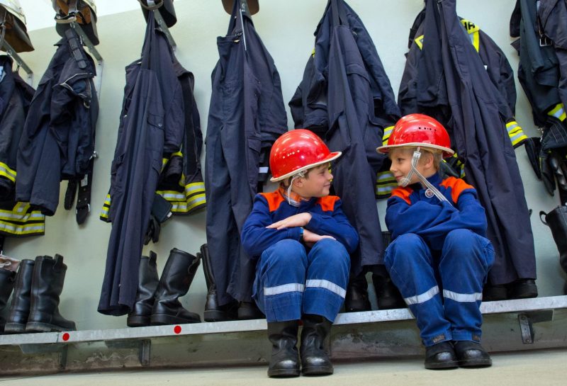 Nachwuchs für die Feuerwehr - zwei Kinder in Feuerwehr-Anzügen sitzen in der Garderobe vor Feuerwehrjacken